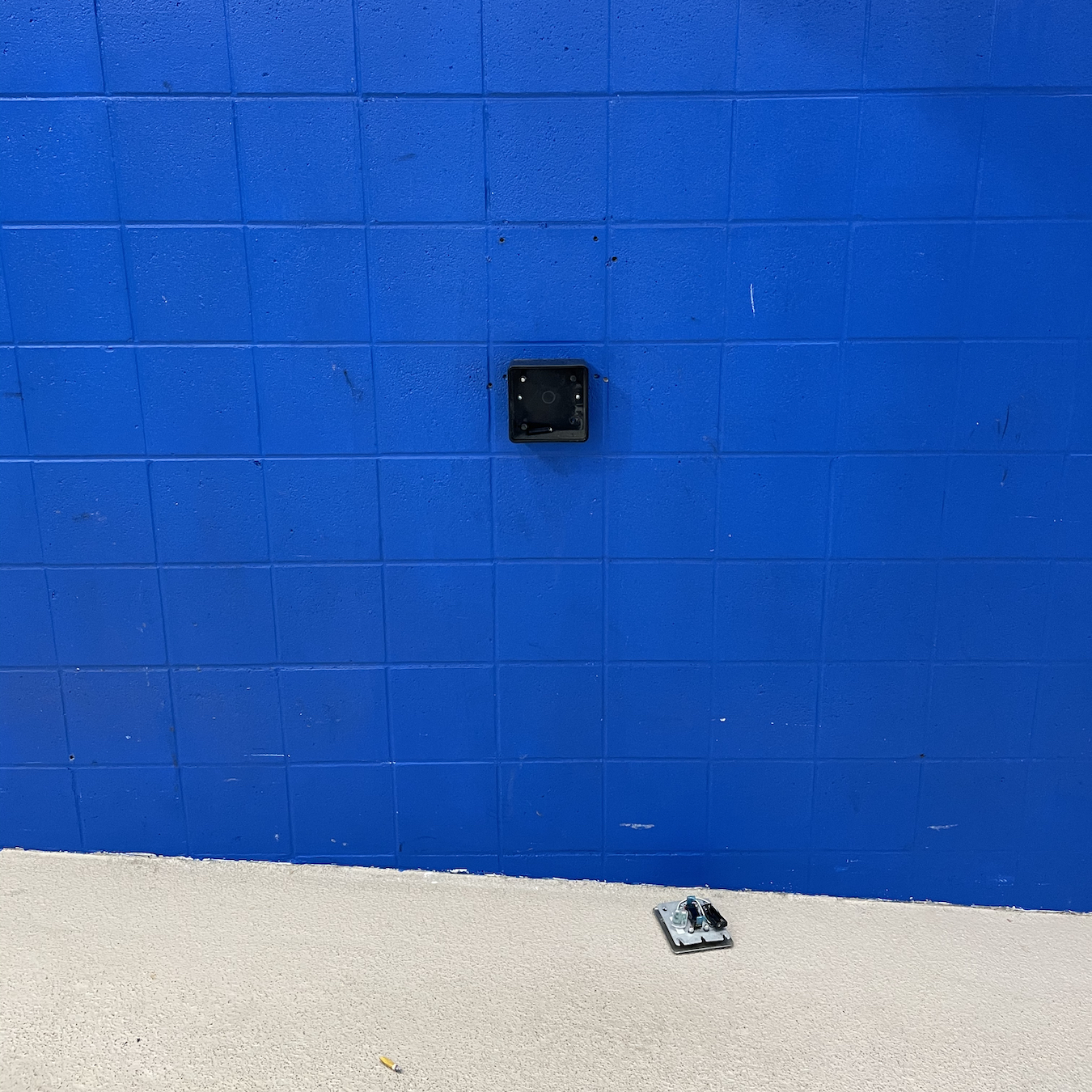 A photo of an accessibility button that would be used to open a door, however the button has fallen outside of the device and onto to the floor, rendering it useless.